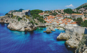 A coastal town with stone buildings and orange rooftops sits beside clear blue water