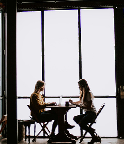 Two women sitting at a table and working on laptops in front of large bright windows.