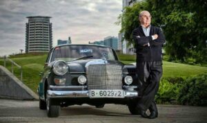 An older man stands with arms crossed, leaning against a classic black Mercedes-Benz car