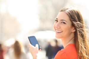 A woman wearing an orange top and smiles while holding a smartphone