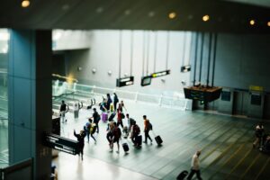 A busy airport terminal with travelers walking and pulling suitcases on a tiled floor, some near escalators and overhead electronic signs.