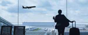 A silhouette of a person holding a cup and rolling a suitcase in front of a large airport window, watching a plane take off under a cloudy sky.
