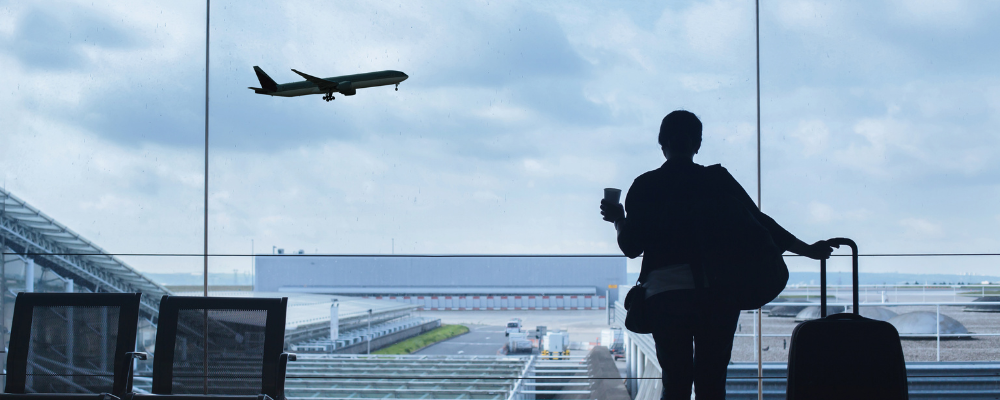 A silhouette of a person holding a cup and rolling a suitcase in front of a large airport window, watching a plane take off under a cloudy sky.