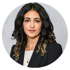 Woman with long curly dark hair and blonde highlights, wearing a black blazer over a white top and a necklace, face intentionally blurred for privacy