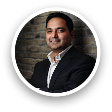 A person wearing a black suit and white dress shirt standing against a brick wall, with the face blurred for privacy, shown in a circular frame with a soft shadow.